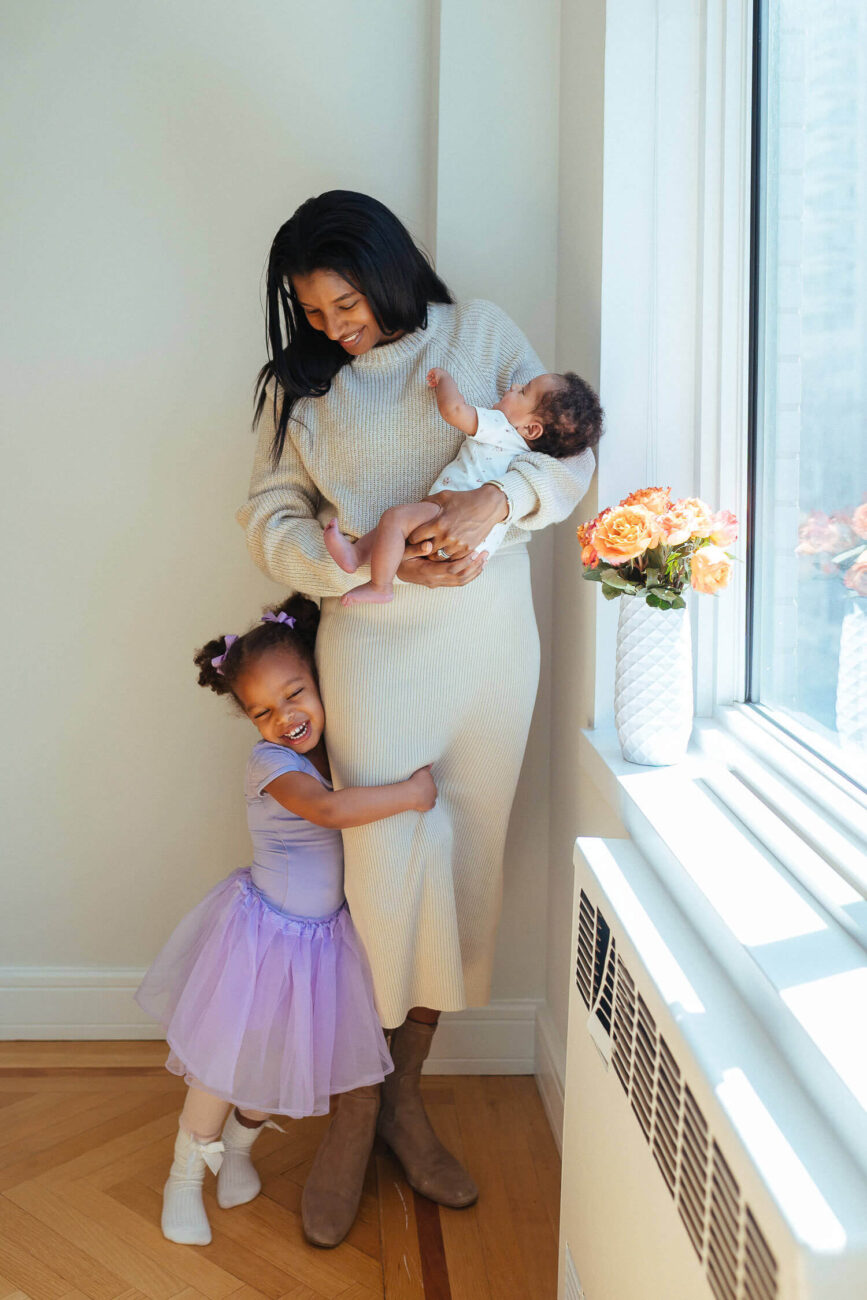 Toodler holding mom's leg, while mom is holding newborn baby in her arms.