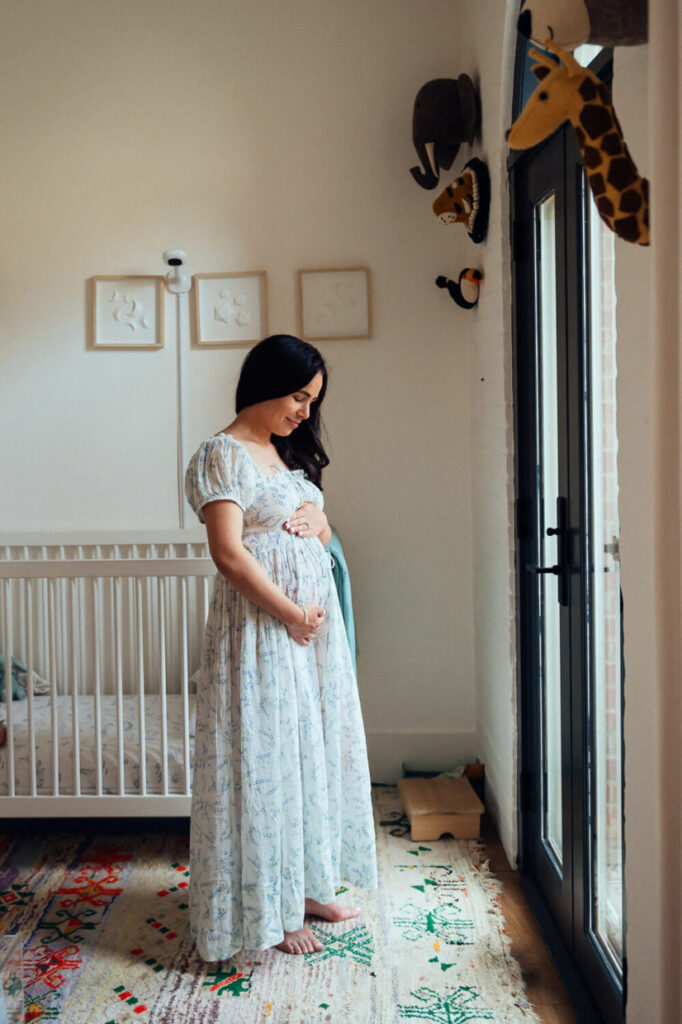 Pregnant woman in a white floral dress standing in a nursery, looking down at her belly.