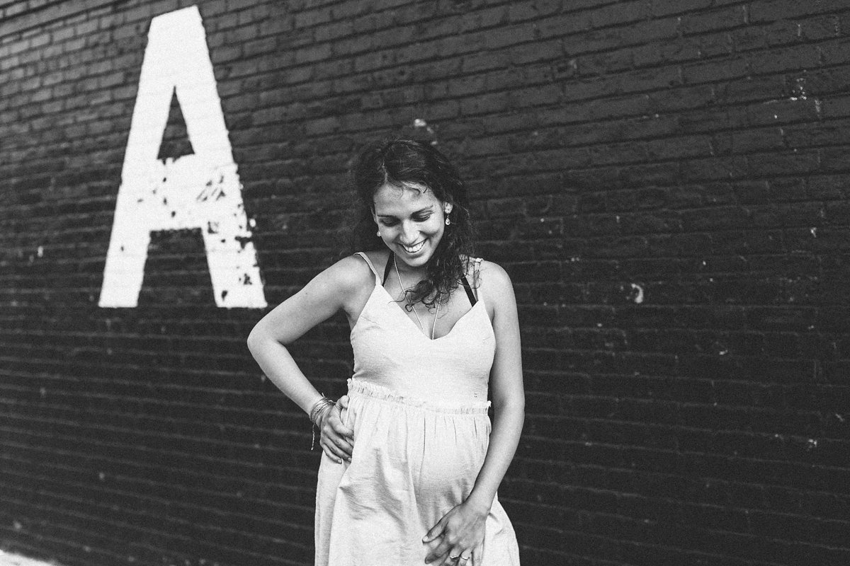 A woman in a stylish light dress stands smiling with her head tilted down, embodying elegant fashion against a black brick wall adorned with a large white letter A. The image captures the timeless allure of monochrome photography.