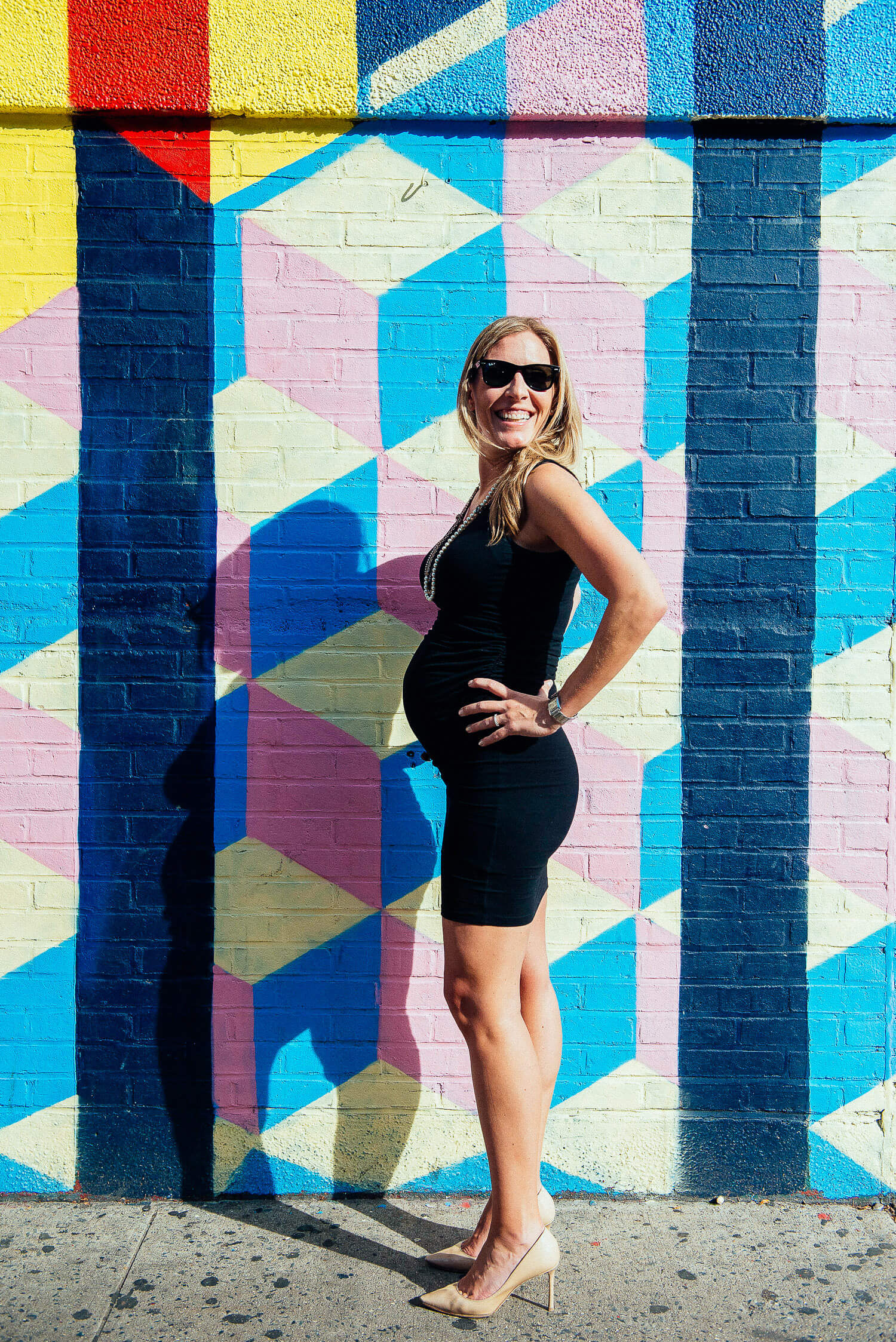 A confident and beautiful mom-to-be wearing a fitted black maternity dress, cool sunglasses, and stylish heels.  Showcasing chic maternity fashion