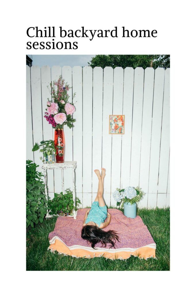 A person lies on their back with feet up against a white fence, relaxing in a serene backyard scene perfect for family sessions. Theyre on a patterned blanket, surrounded by flowers in vases on stands. The text above reads Chill backyard home sessions.