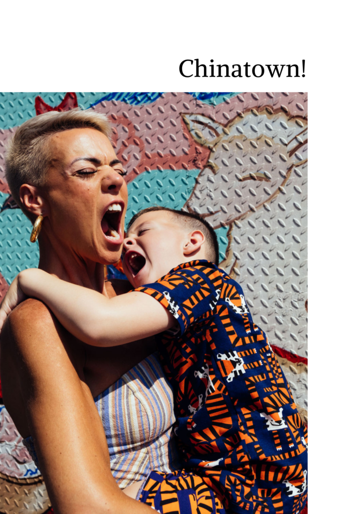 A woman with short hair holds a young boy in her arms, both playfully shouting or laughing. The boy wears a colorful patterned shirt. Behind them, a textured wall boasts whimsical illustrations and Chinatown! stands out prominently. Capture the joy of Unique Family Sessions 2025!.