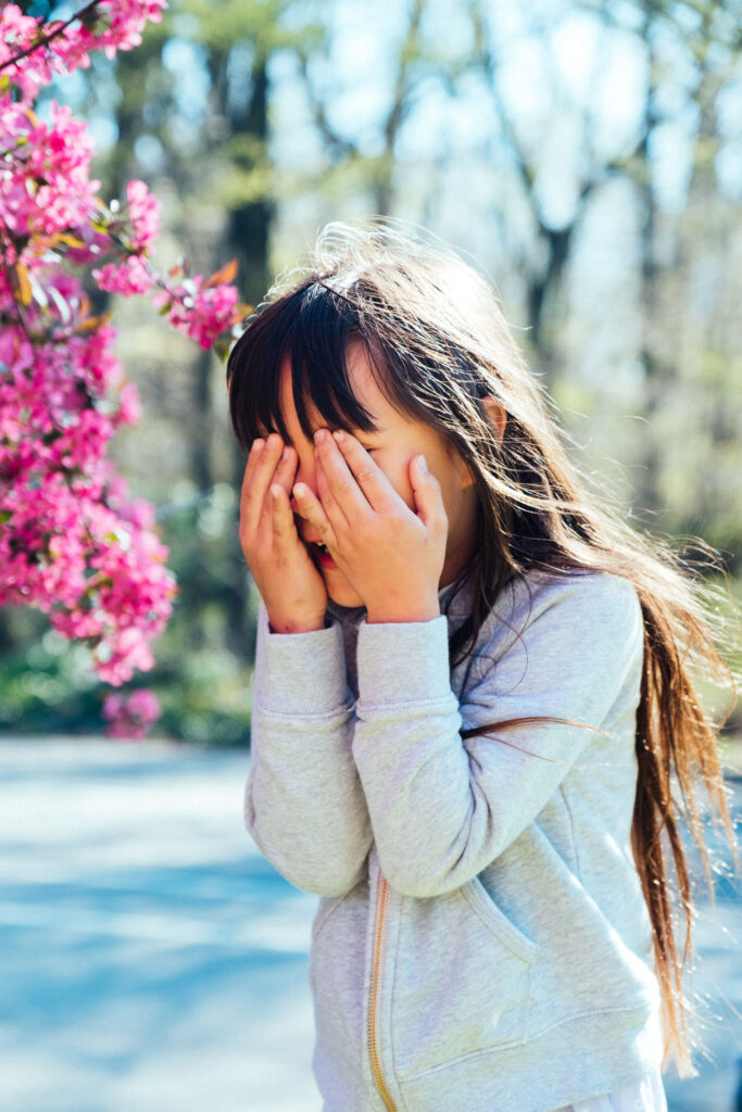 A young girl with long hair and wearing a gray hoodie covers her face with her hands. She stands outdoors near a branch of bright pink cherry blossoms, reminiscent of the best locations in NY, with blurry trees in the background on a sunny day.