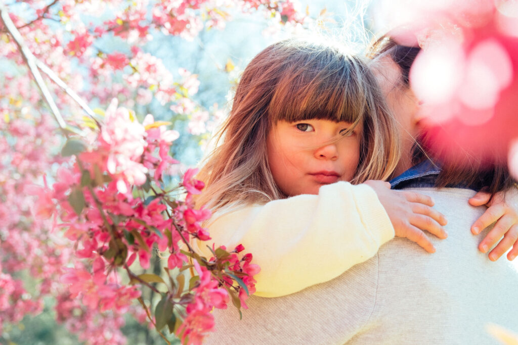 A young girl with long hair is held by someone in a light top, surrounded by cherry blossoms on a sunny day. She calmly gazes at the camera amidst the vibrant blooms, capturing one of NYs best locations for enjoying these cheerful flowers.