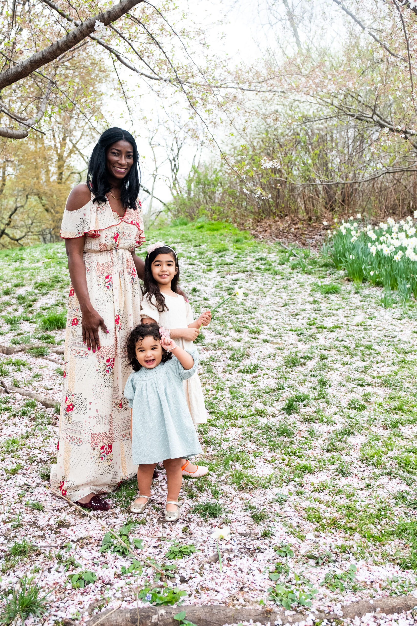 A woman stands with two young girls on a path covered in pink petals, surrounded by blooming daffodils and cherry blossoms in a picturesque NY park. The woman wears a floral dress, and the girls don pastel ones, all smiling at the camera. Its one of the best locations for capturing springs beauty.