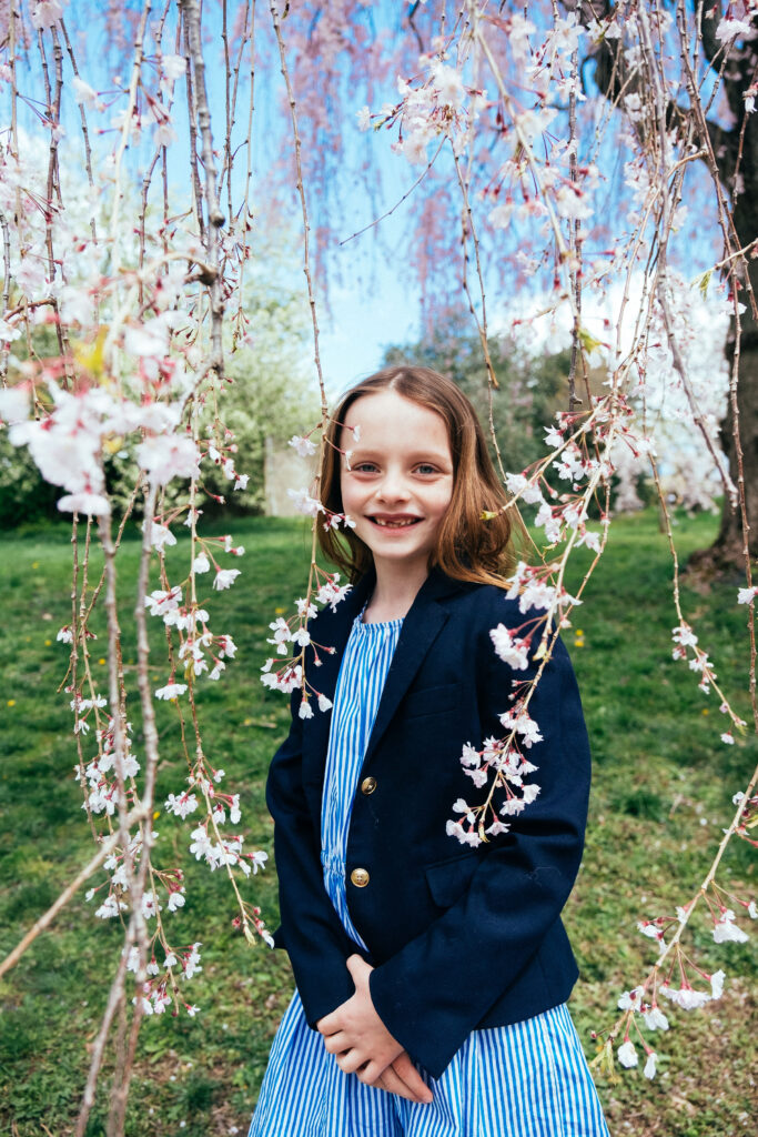 A young girl in a blue and white striped dress with a black blazer stands smiling among the cherry blossoms, embracing one of NYs best locations. Shes outdoors on a grassy lawn, framed by trees under the clear blue sky.