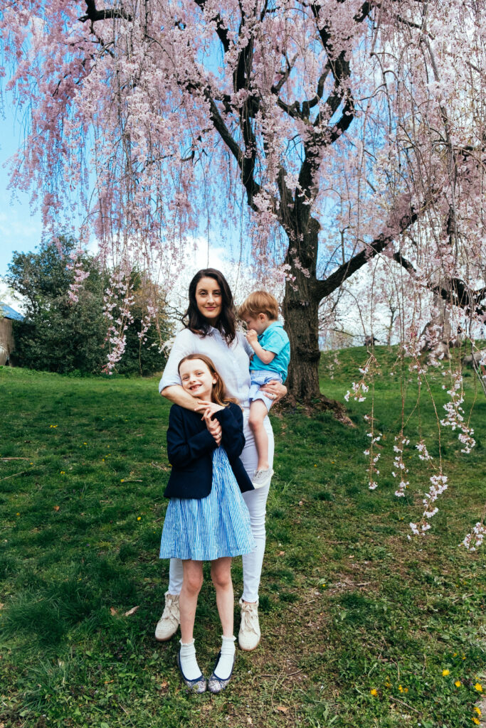 A woman stands smiling under the cherry blossoms in NY, embracing her two children. The girl wears a striped dress and beams, while the young boy in a light blue outfit is held by the woman. They are on a grassy lawn with scattered flowers.